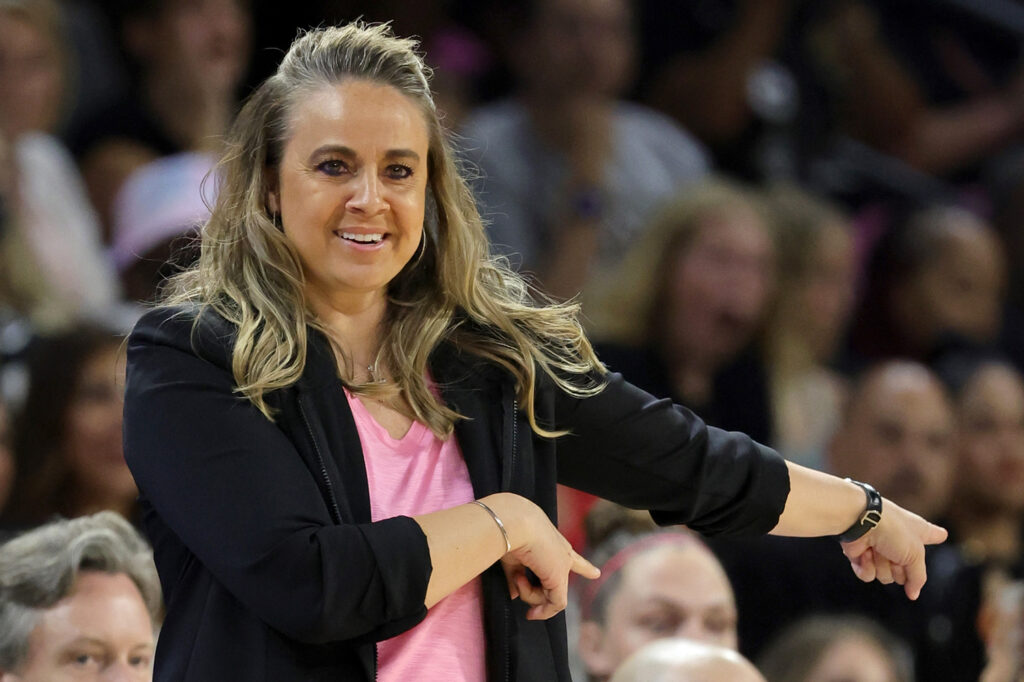 Becky Hammon smiles in front of spectators while gesturing to her left.