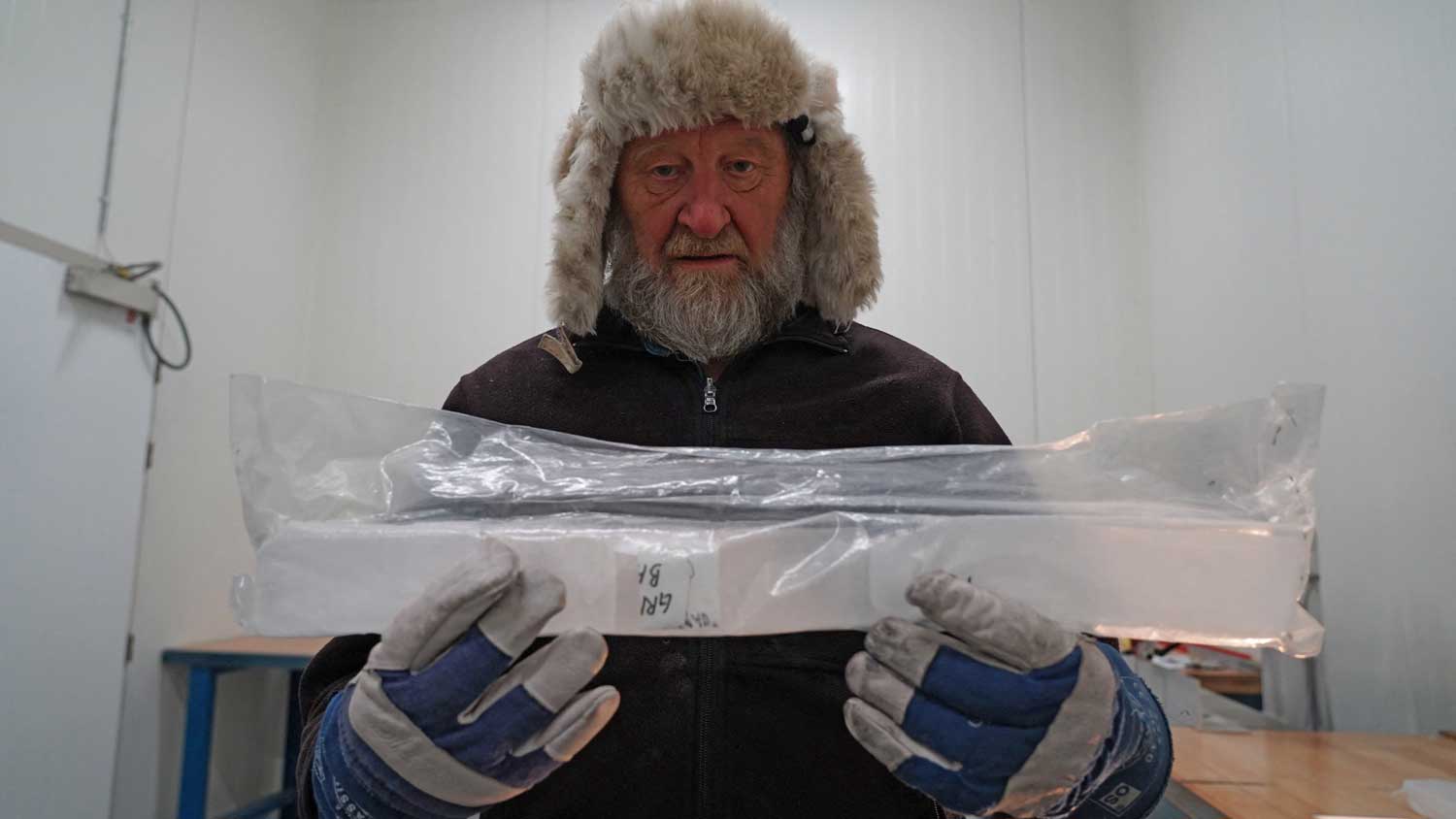 A man wearing a fur hat and gloves holds out a core of ice in a labeled plastic bag.