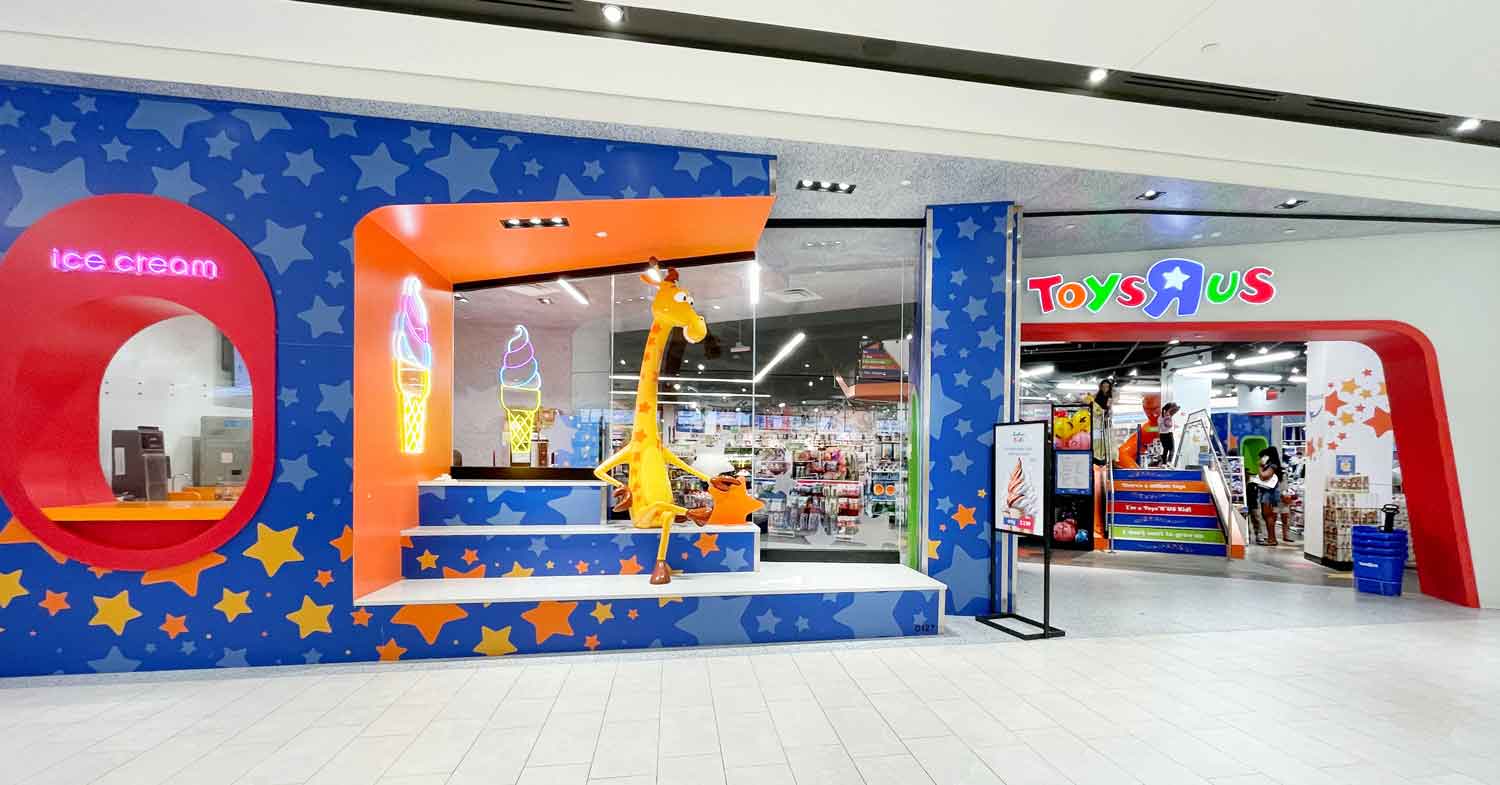 Storefront of a mall Toys R Us with windows showing Geoffrey the giraffe and an ice cream area with the words ice cream and a lit up ice cream cone.