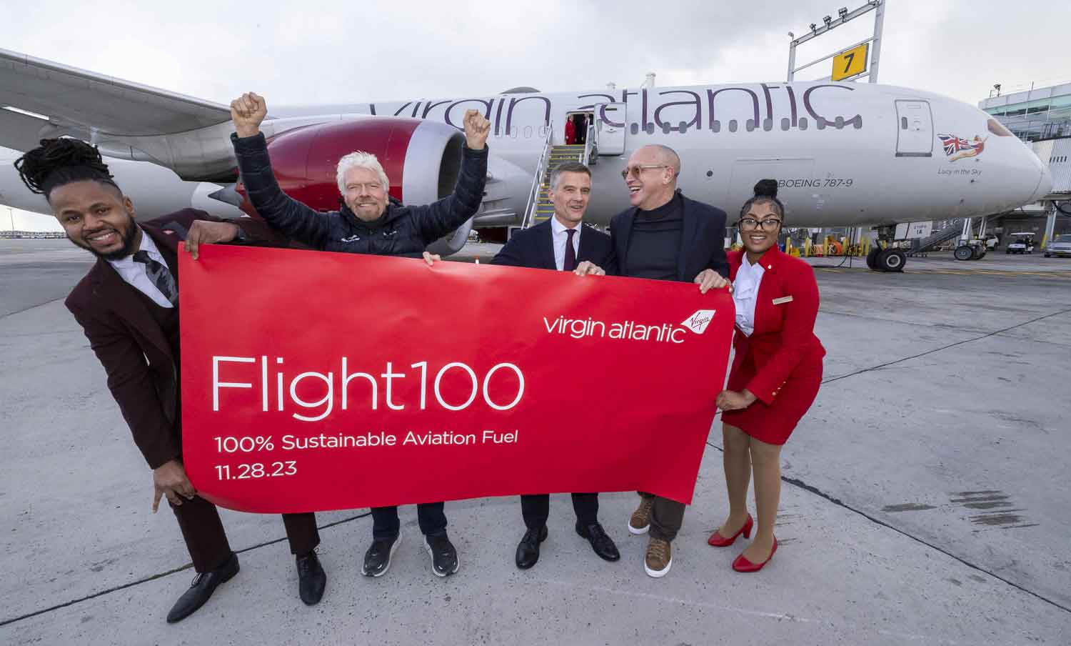 Richard Branson, three other men, and one woman pose in front of a plane holding a banner that says Flight100.