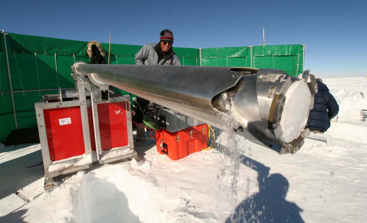 A person uses a large metal cylinder to extra a core of ice from the ground.