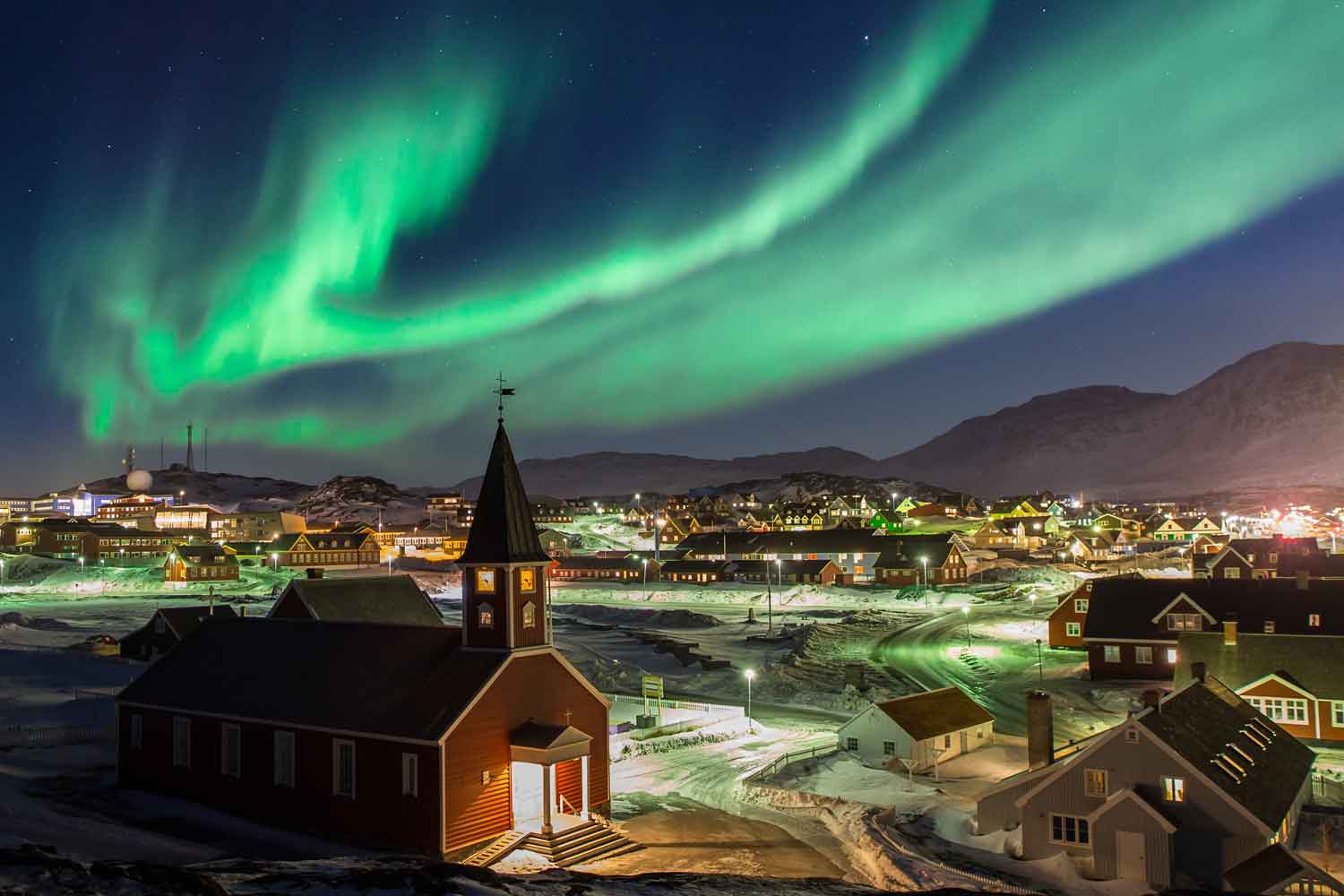 The Northern lights are in the sky over a city of homes and other small buildings.