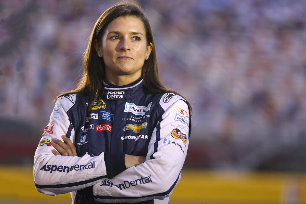 Danica Patrick stands with her arms folded while wearing a jacket on which several sponsor names appear.