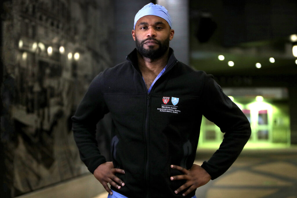 Myron Rolle poses while wearing a black zip up fleece over blue hospital scrubs.