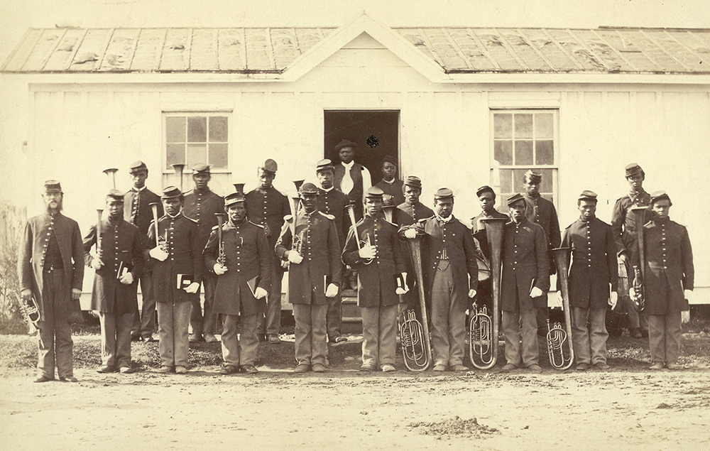 Black Civil War soldiers in uniform pose in front of a building holding musical instruments.