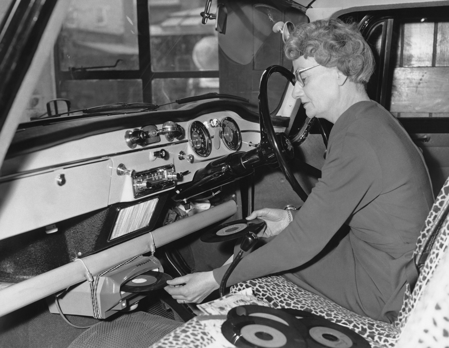 A woman sits at the steering wheel on an old car and puts a 45 record into a below-dashboard record player.