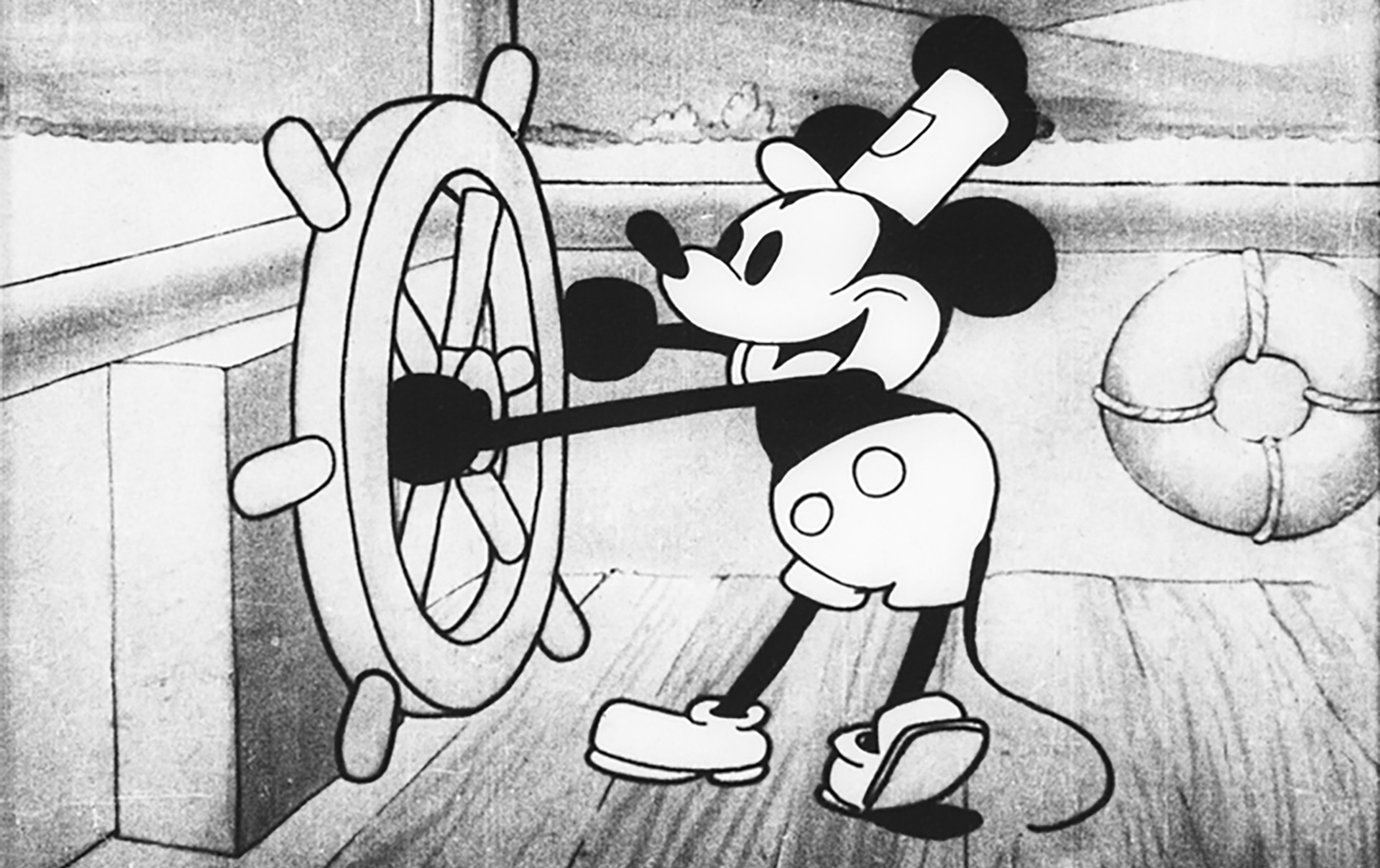 A black and white Mickey Mouse wears a hat and holds onto the wheel of a boat.