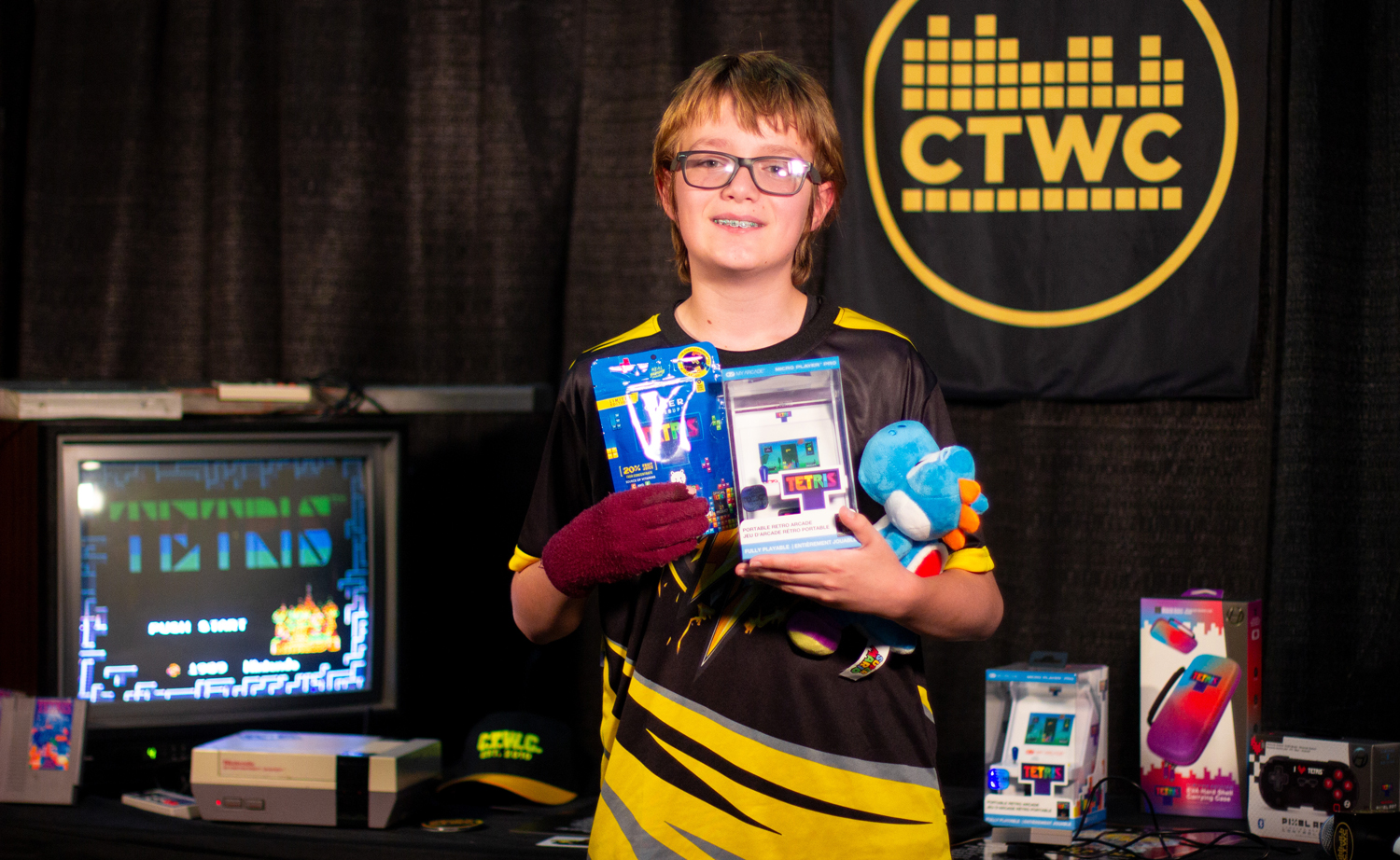 A young teen poses in front of a video game system and holds Tetris accessories in his arms.