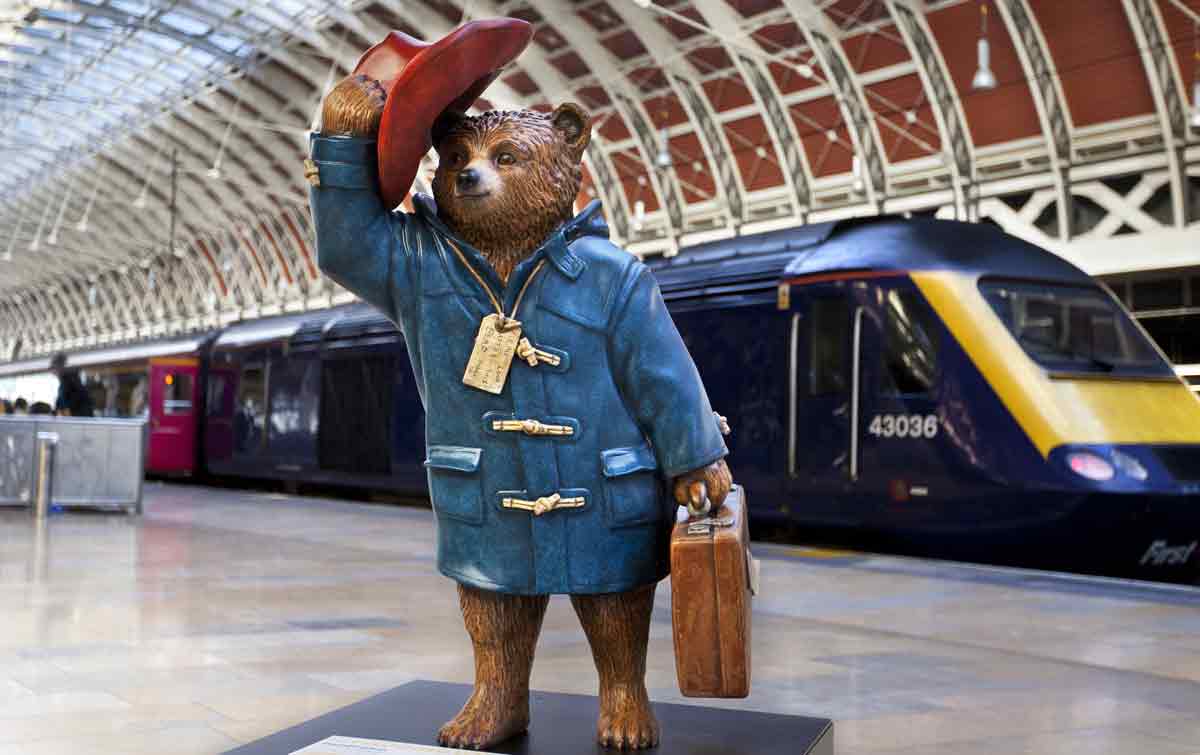 A statue of Paddington wearing a blue coat and tipping his red hat stands on a train platform.