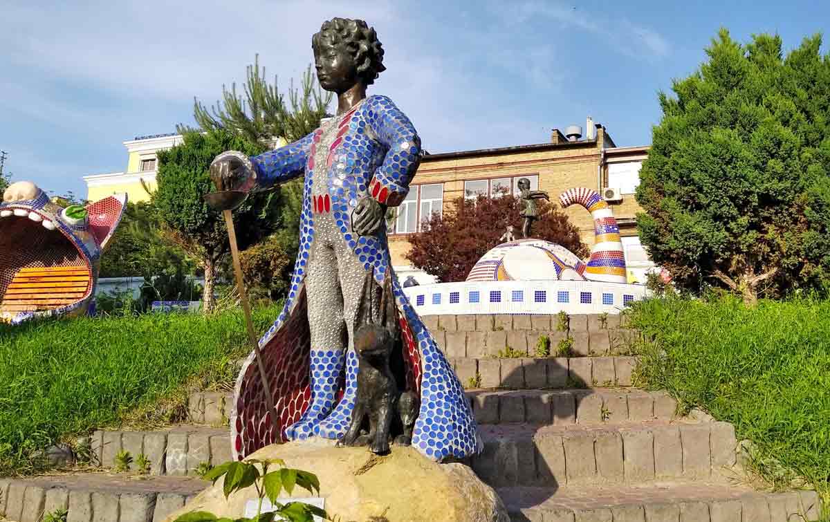 Statue of the Little Prince wearing a mosaic coat, holding a sword, and standing next to a fox.