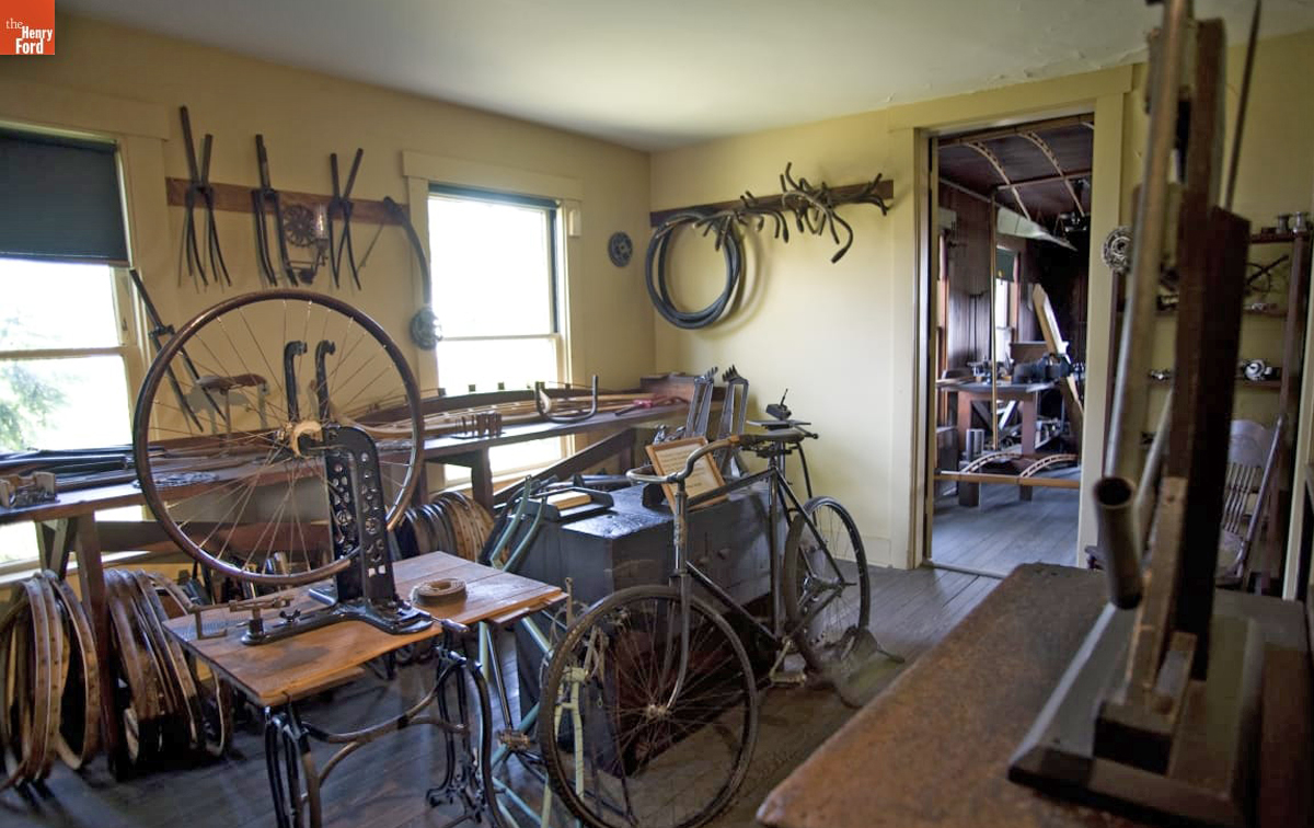A bicycle and bicycle parts sit in and around 19th century storage and equipment in a room.