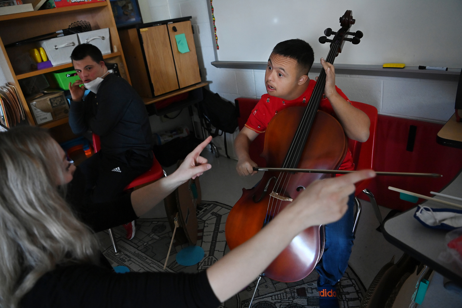A high school student plays the cello and another sits in a chair as both look at Annie Ray as she provides instruction.