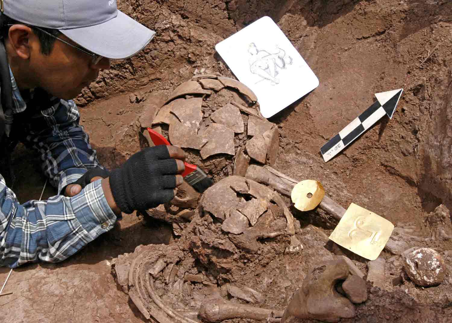 A man lies on his stomach and applies a brush to an object at an archaeological site.