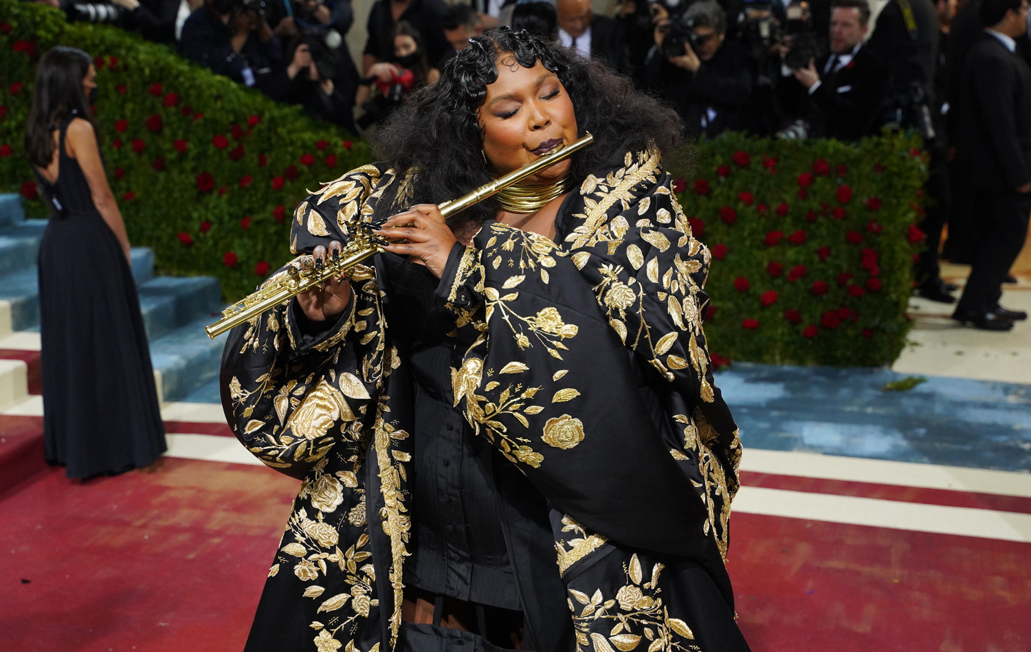 Lizzo is dressed in formal wear as she plays a flute on a red carpet in front of a crowd of photographers.