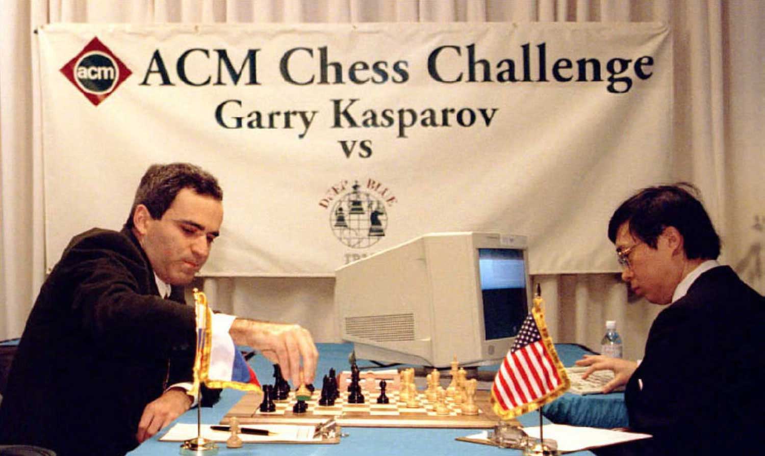 A man moves a piece on a chess board as another man watches and types into a computer.
