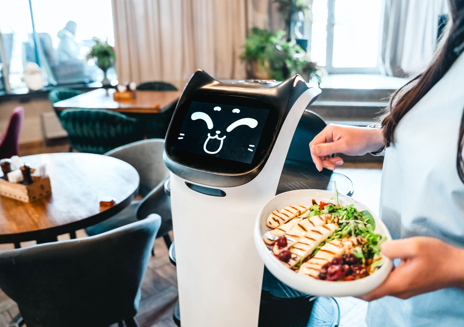 A robot with a dog’s face is next to a restaurant server with a plate of chicken and vegetables.