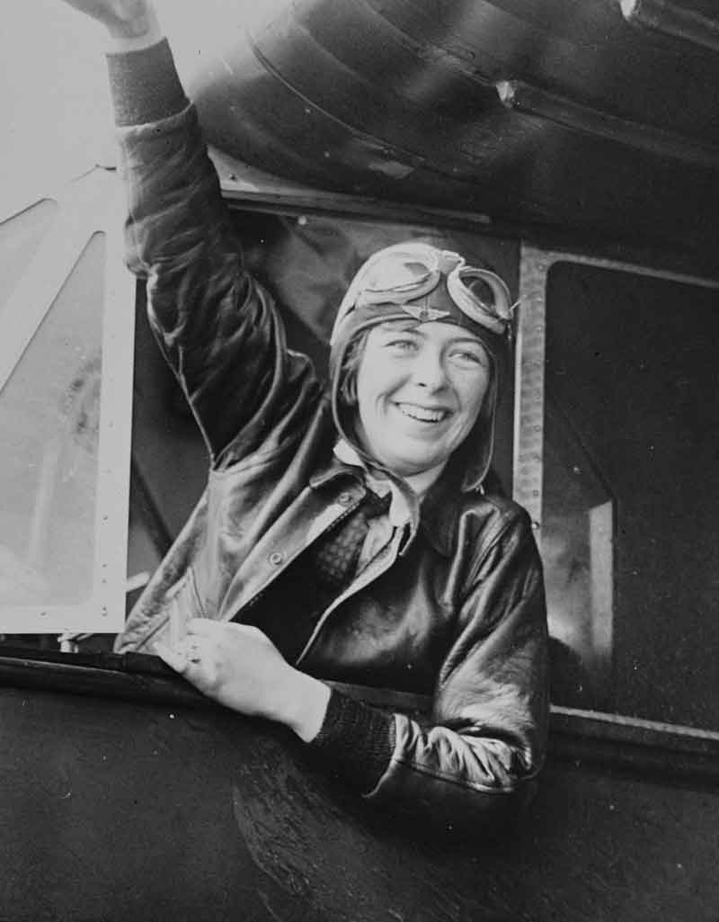 Elinor Smith wears an aviator jacket with goggles on her head as she smiles and waves from an airplane cockpit.