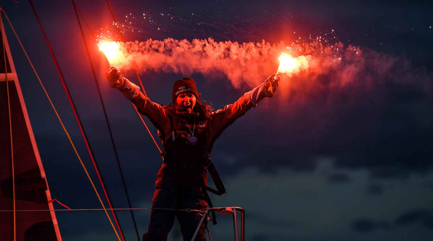 A woman stands on the prow of a boat and smiles as she holds out two flares.