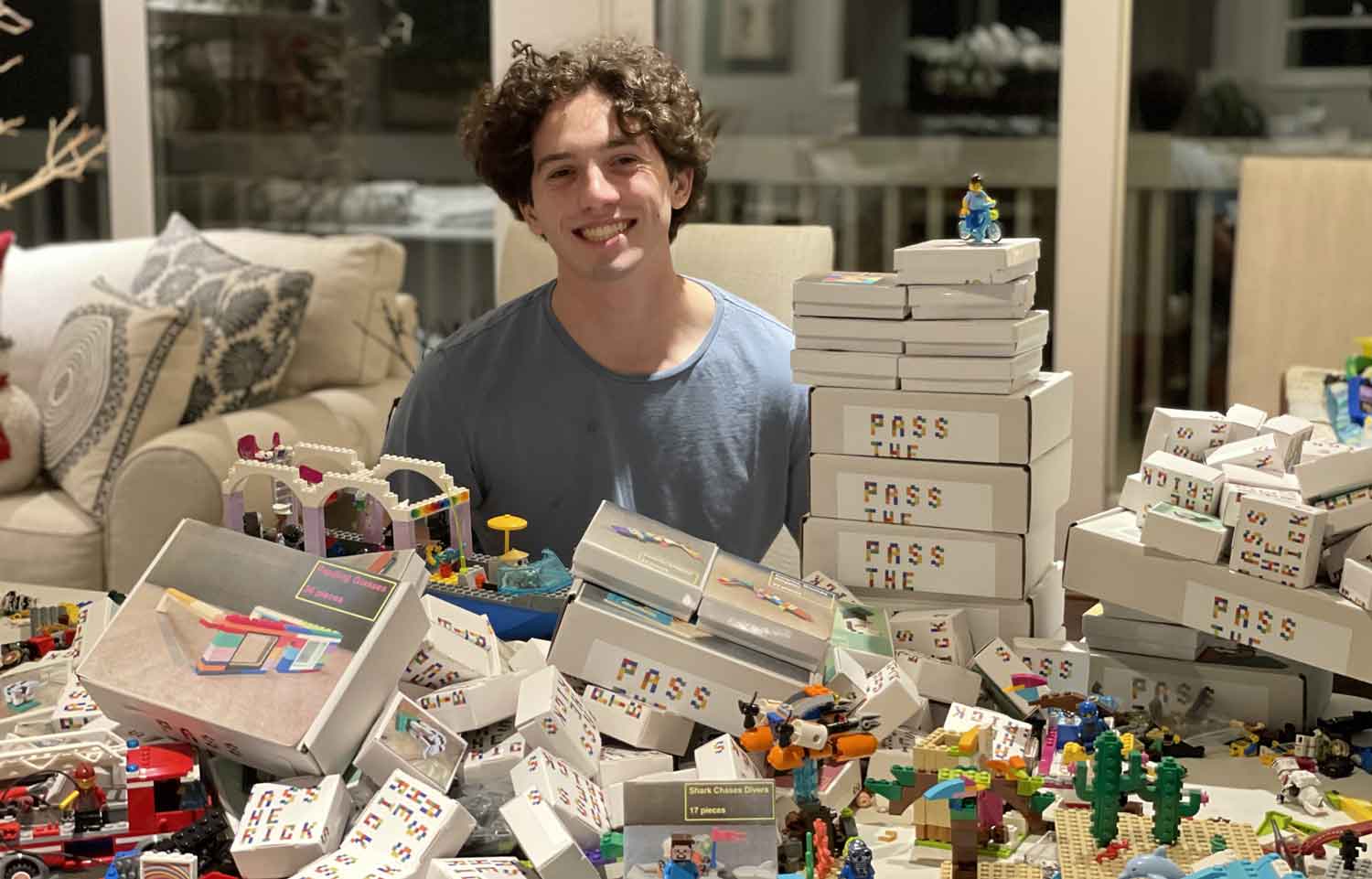 A teen smiles behind a table with many LEGO bricks and white boxes labeled Pass the Bricks.