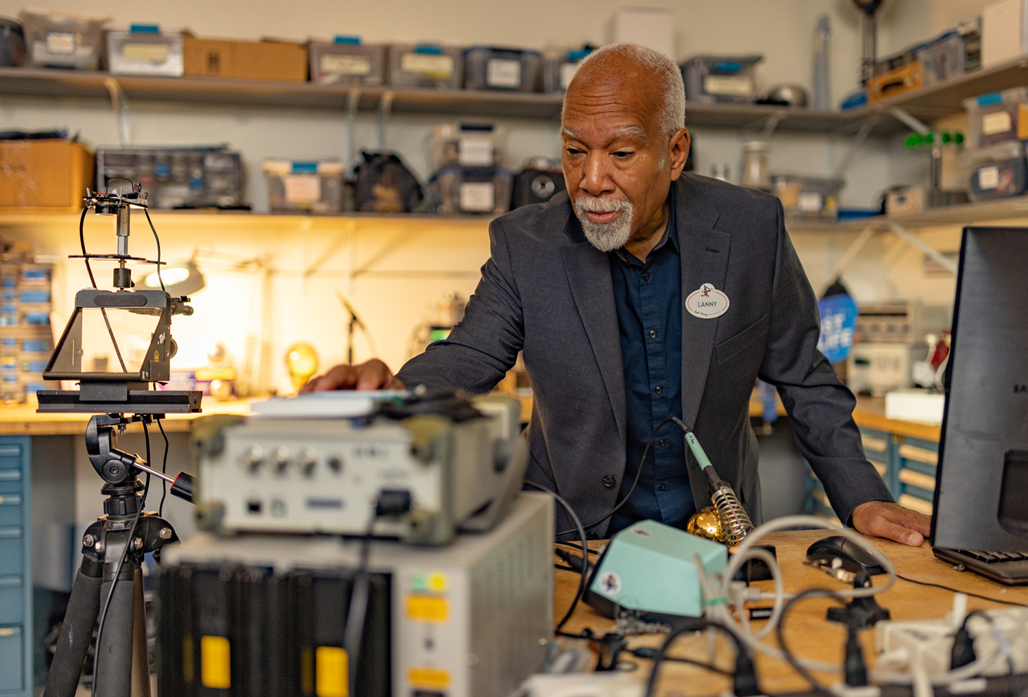 Lanny Smoot leans down and manipulates a device while surrounded by many computers and other types of technology.