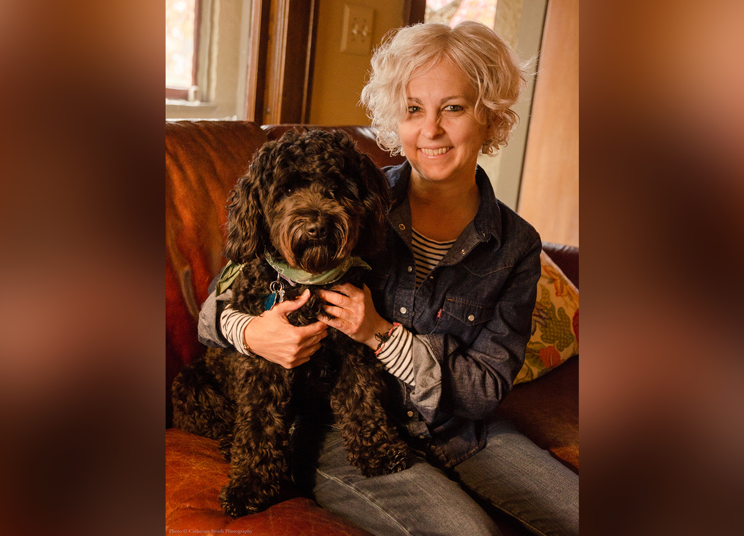 Kate DiCamillo sits on a couch with her arm around a black dog with curly fur.