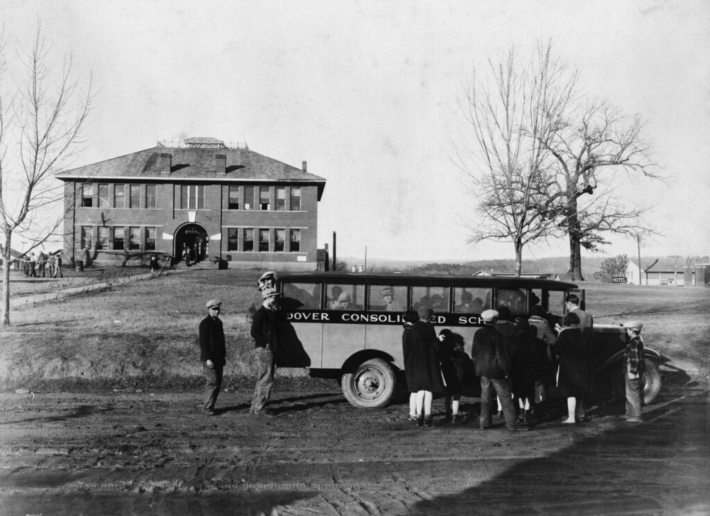 Adults in 1930s clothing stand around a 1930s school bus in front of a school in a rural area.