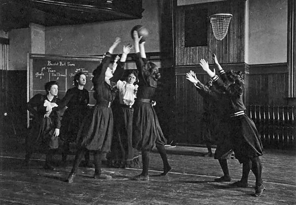 A group of young women in knee-length uniforms play basketball with a female coach watching.