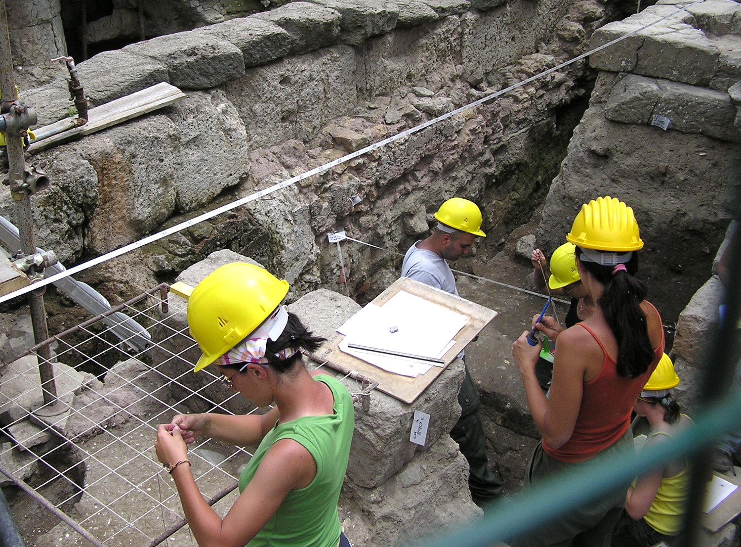 Five archaeologists wear yellow hard hats as they work among ancient ruins.