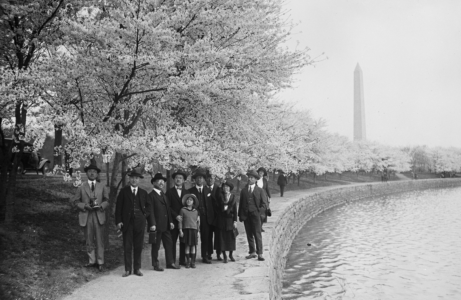 A group of men, women, and one child in 1920s clothing stand under cherry blossoms with the Washington Monument in the background.