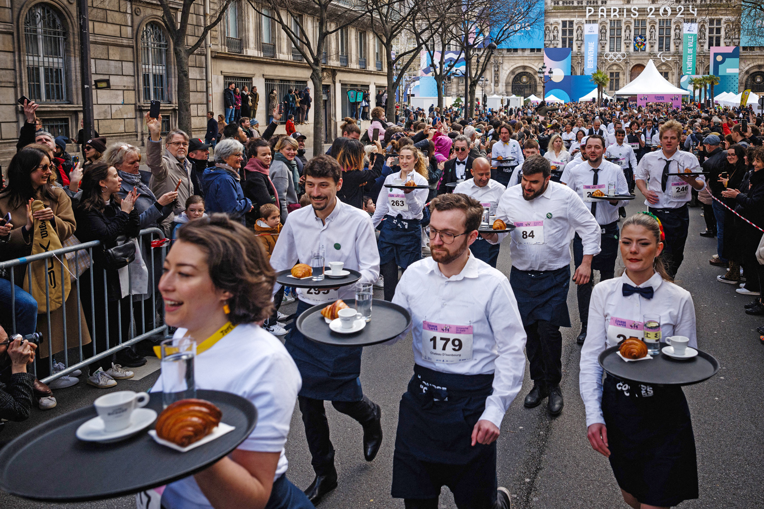 A group of servers in white shirts carry trays with croissants, water, and coffee as spectators watch from the sidelines.