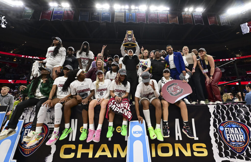 A group of young women pose in t-shirts that read “Champions” and a banner that reads “Final Four National Champions” as their coast holds up a trophy.