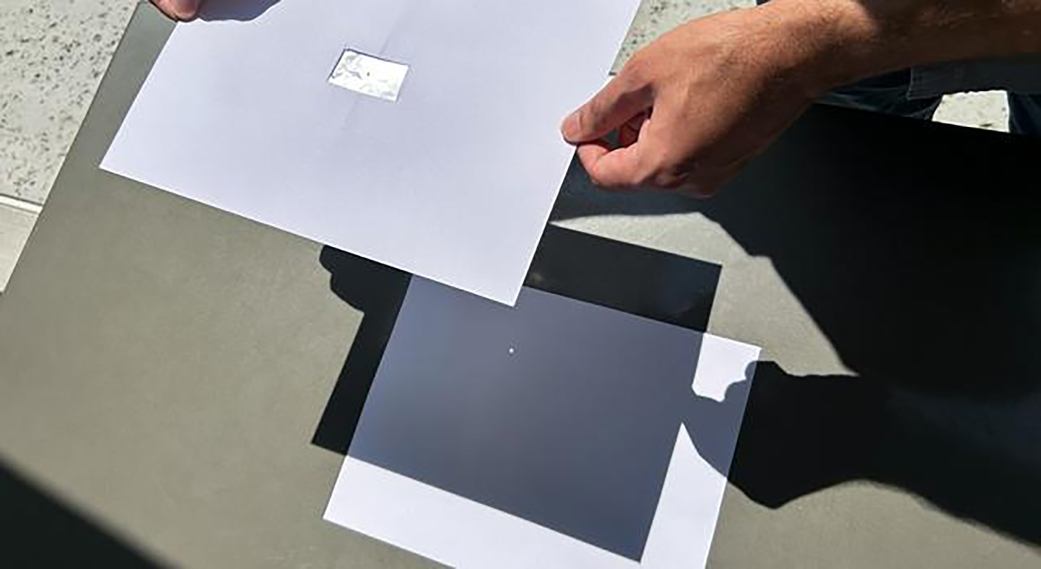 Two hands hold a piece of paper with a rectangle of foil in the center over another piece of paper in sunlight.