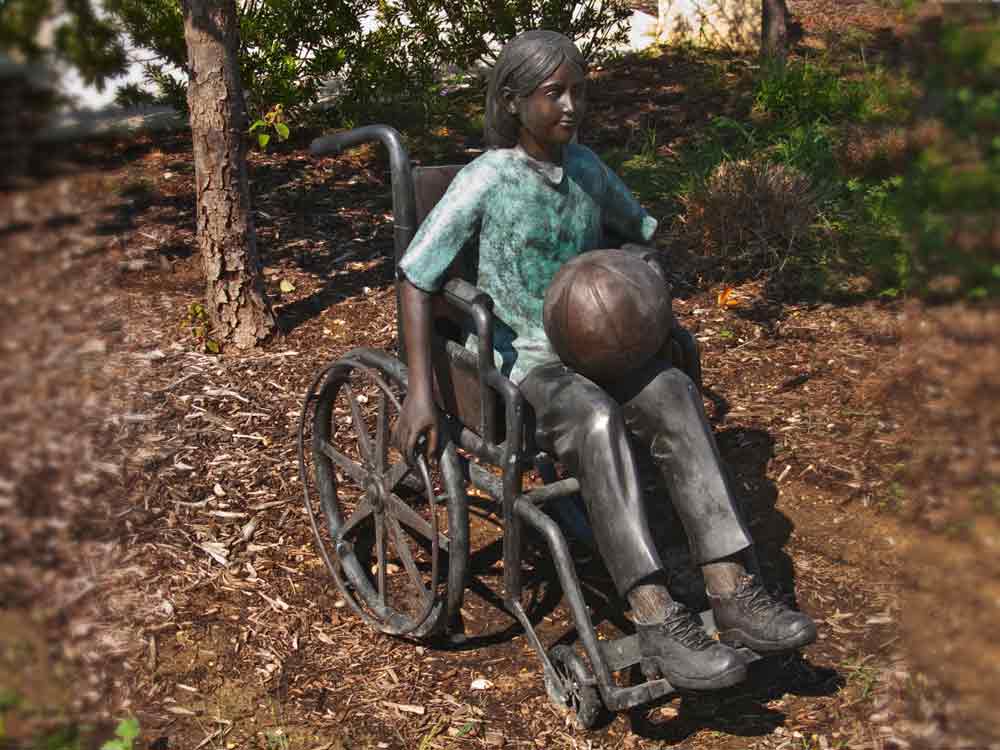 A metal sculpture of a child sitting in a wheelchair with a ball on her lap.