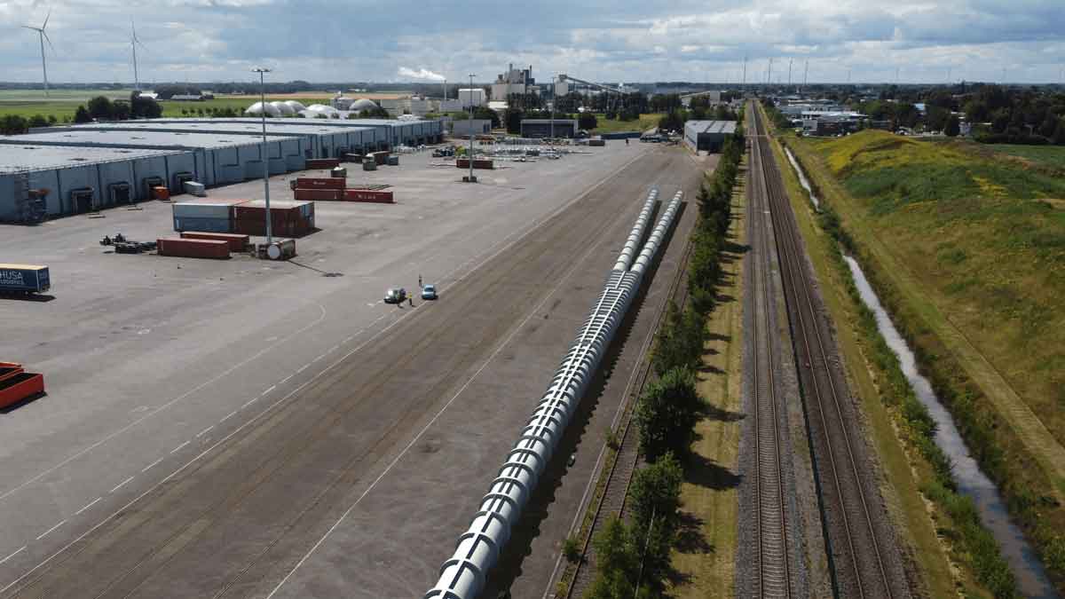 A white tube runs alongside train tracks and next to a large complex of rectangular buildings.