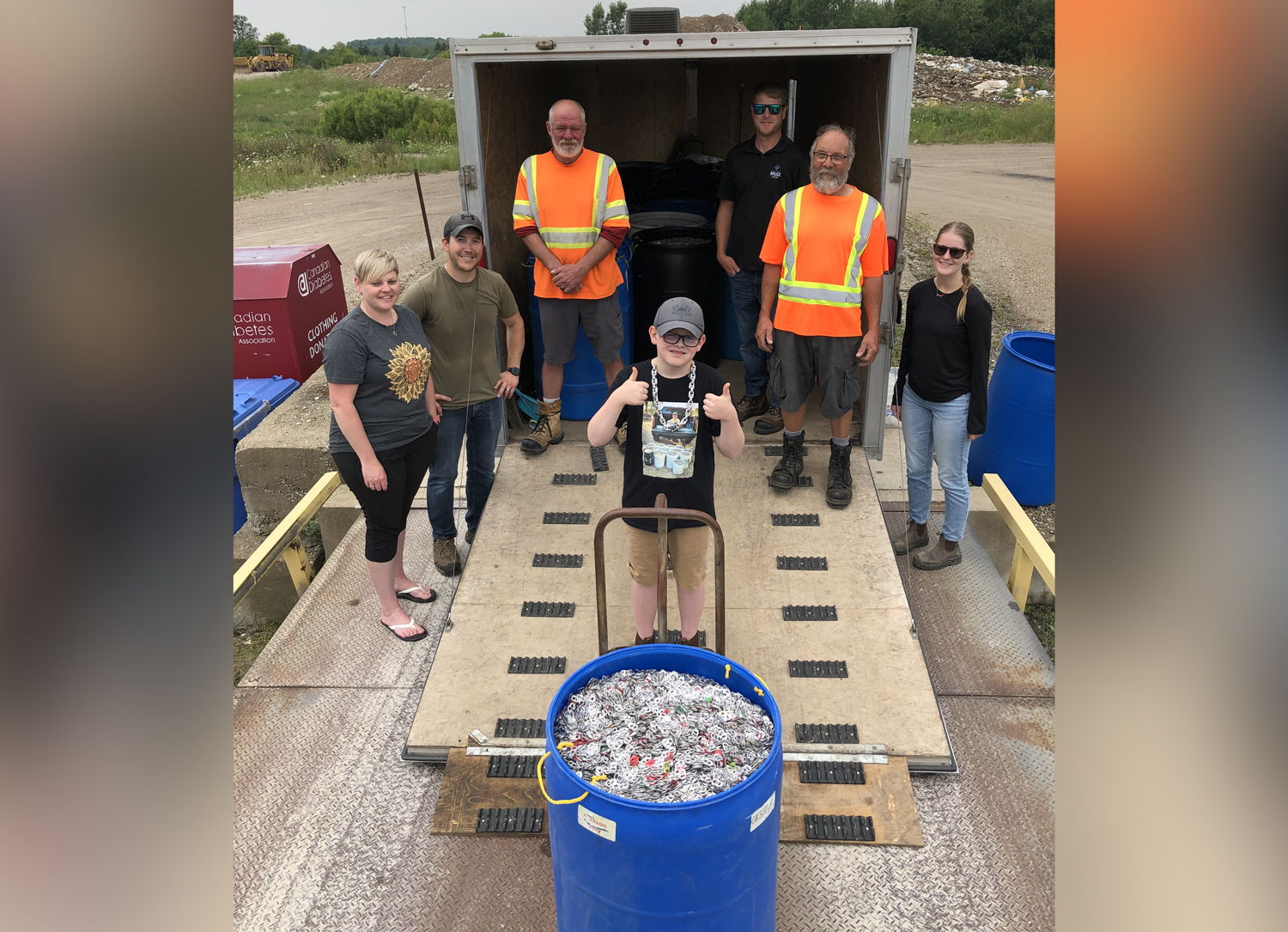 A child gives two thumbs up on the back of a truck with a barrel of can tabs, his parents, and four other adults.