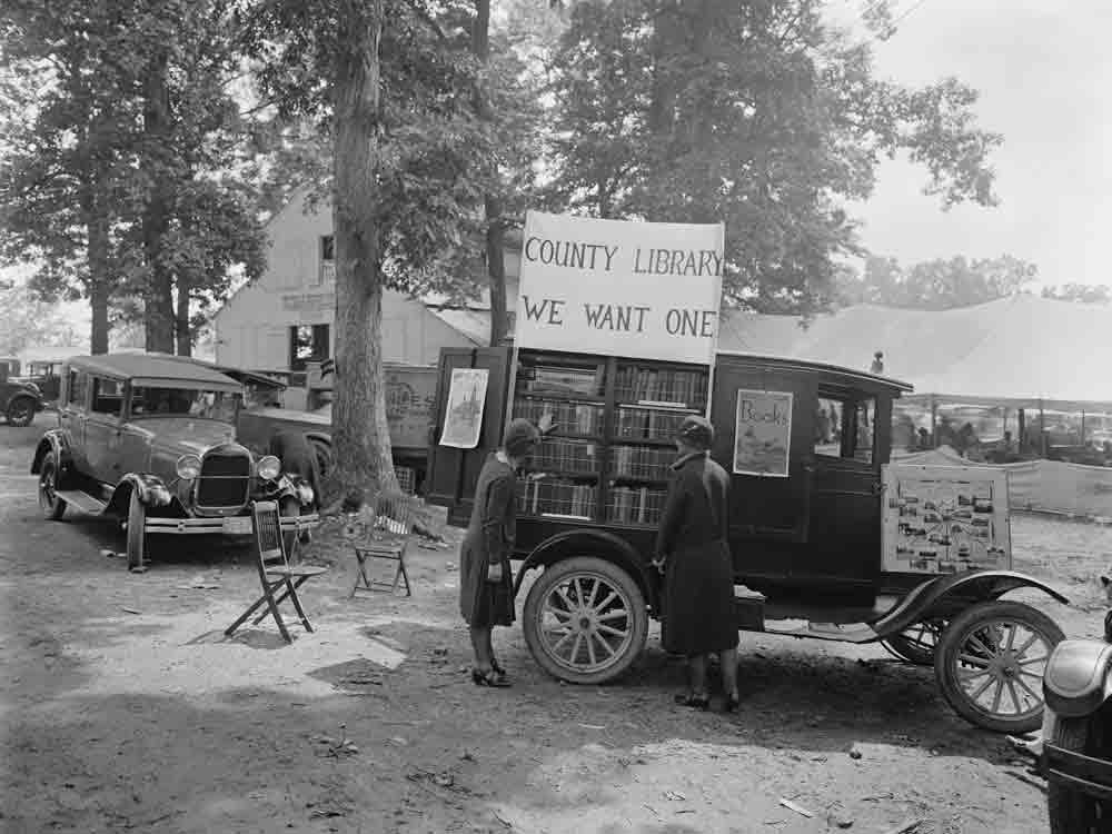 Two women in 1920s clothing at a 1920s car that has been turned into a bookmobile and point at books.