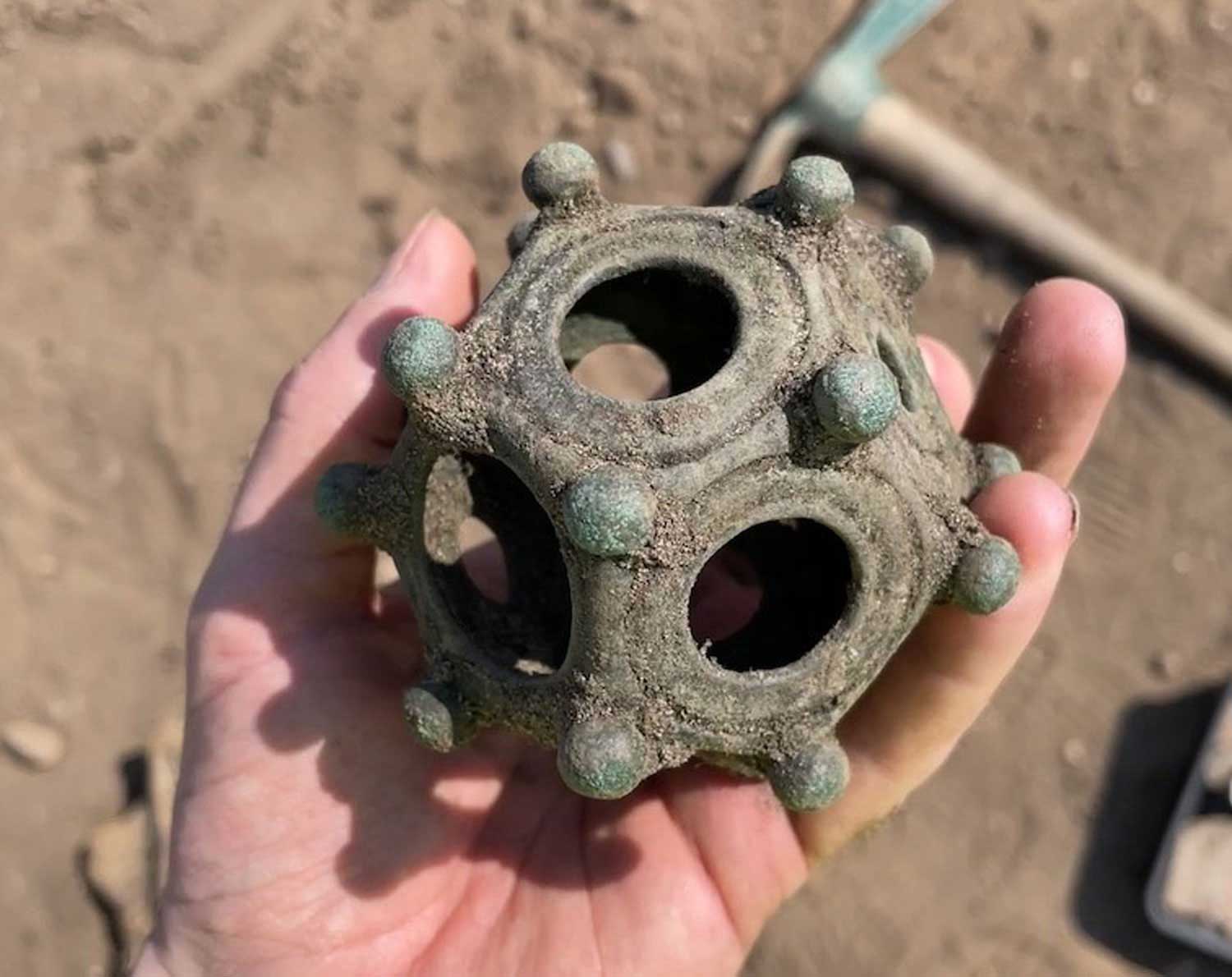 A metal dodecahedron is held in a hand at an archaeological site.