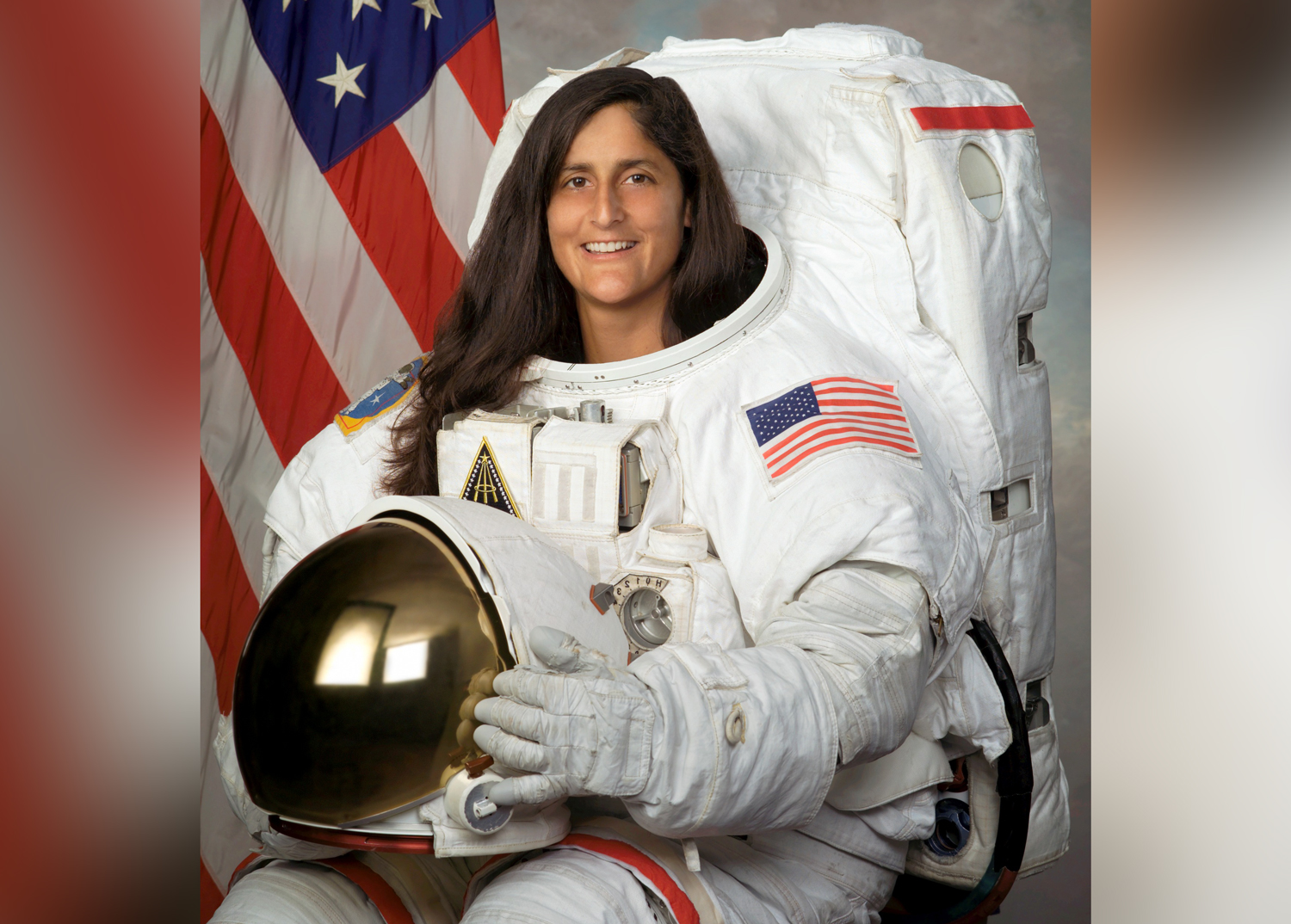 Sunita Williams poses in a spacesuit in front of a U S flag.