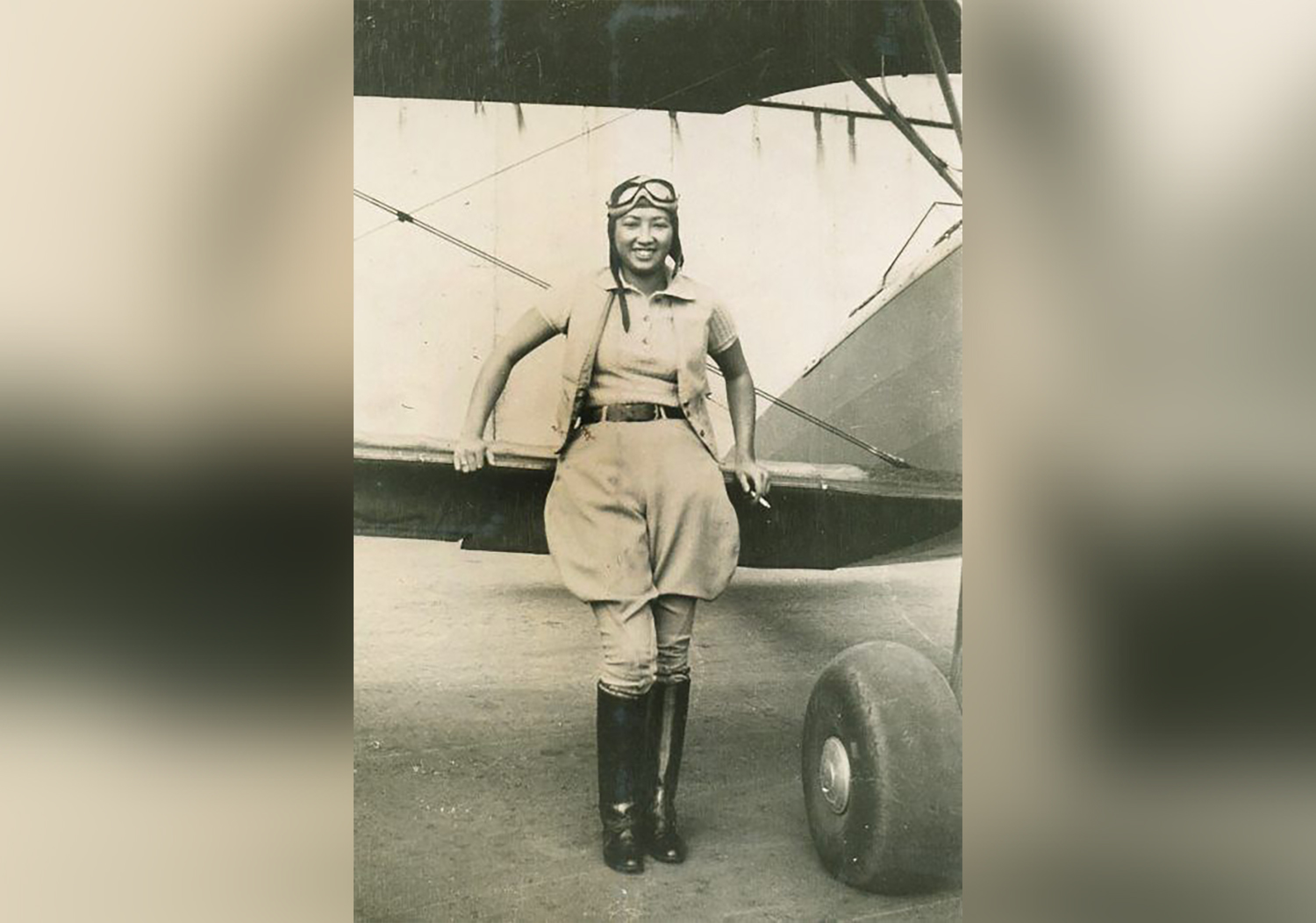 Hazel Ying Lee stands in front of a small plane and wears 1930s aviation clothing and goggles.
