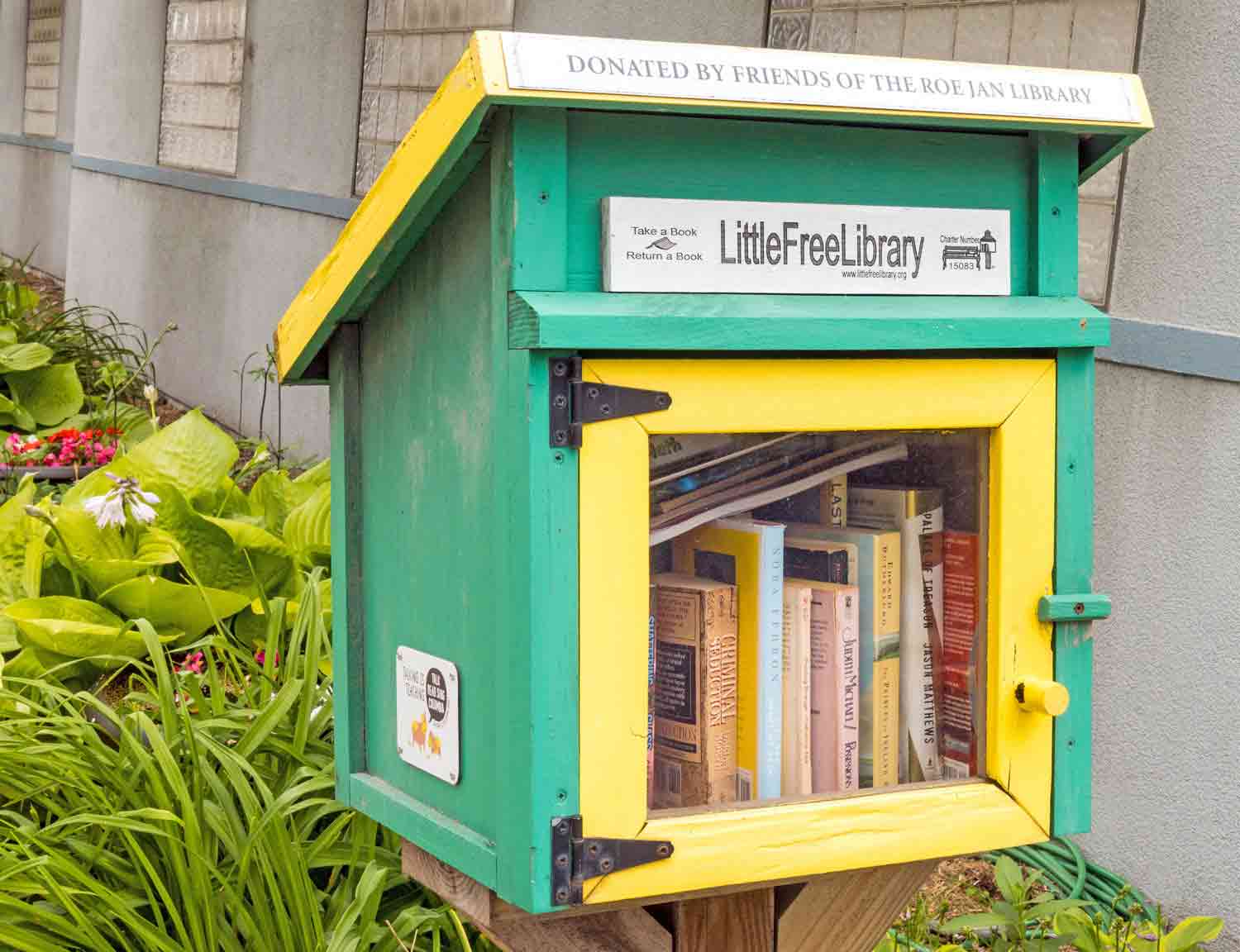 A small green and yellow shed on a post is labeled Little Free Library and is filled with books