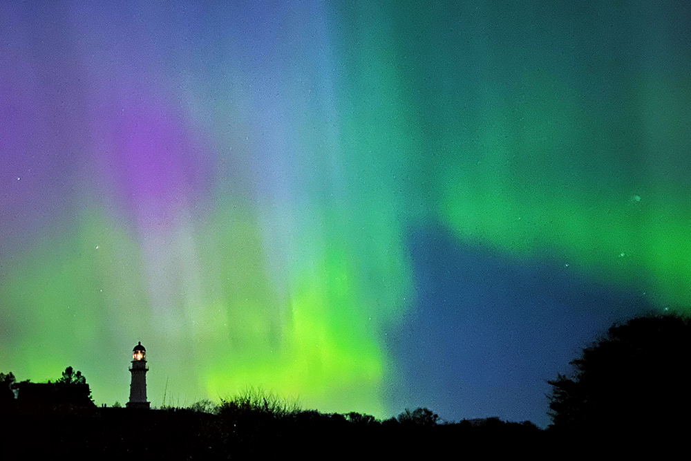Streaks of yellow, green, and purple in the night sky above a lighthouse