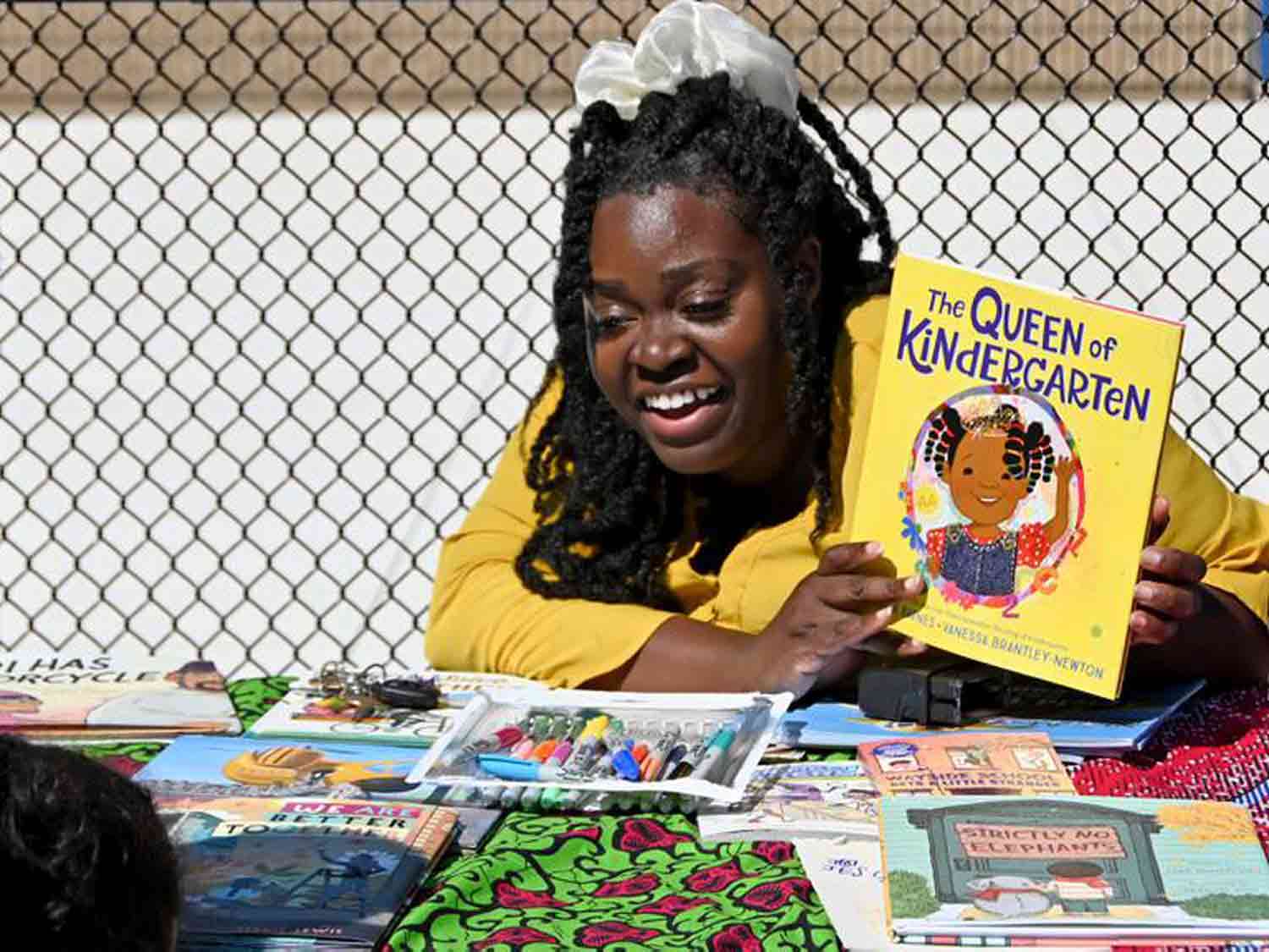 Storybook Maze smiles at a child while sitting behind a table of books and holding up a book called Queen of Kindergarten.