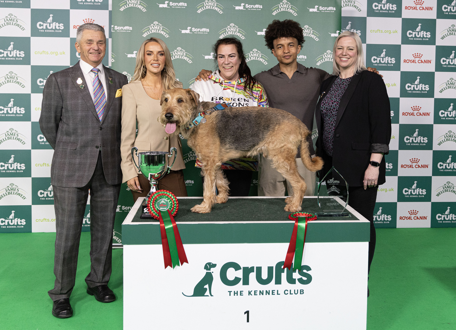 Five people pose behind a black and tan dog that stands on a table with ribbons and trophies.