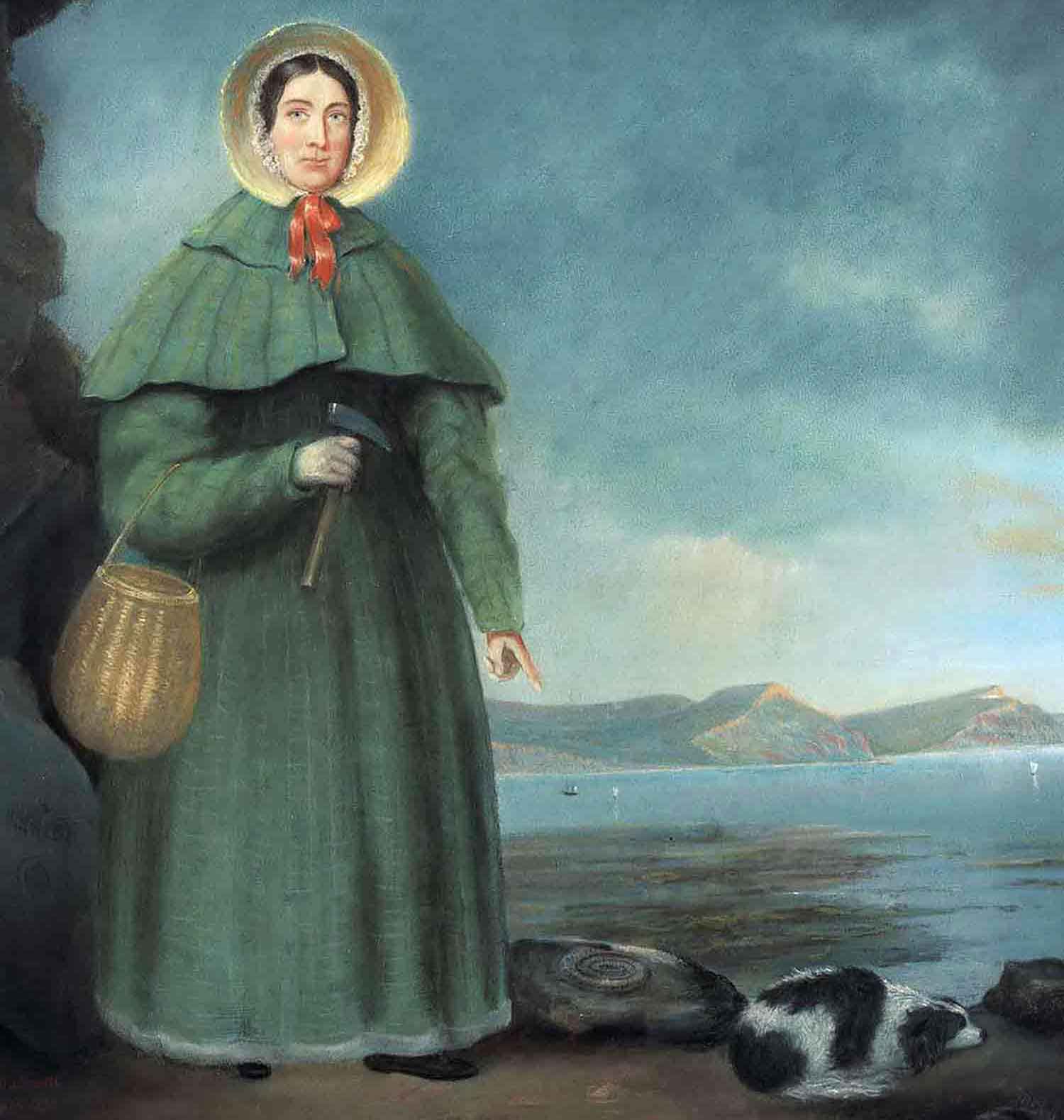 Mary Anning holds a digging tool and points to a fossil as she stands next to her dog.