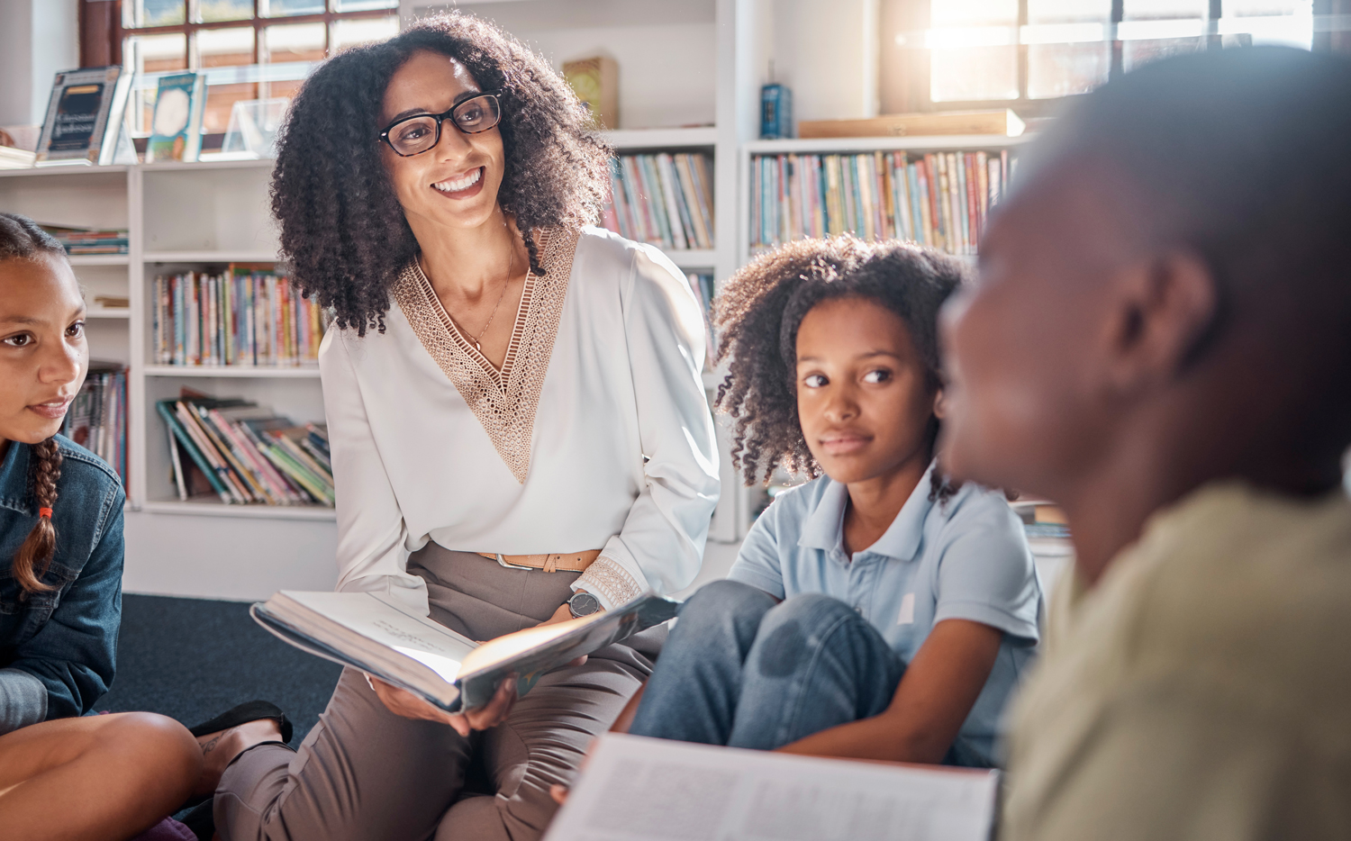 A librarian and three kids sit on the floor of a library, reading a book.