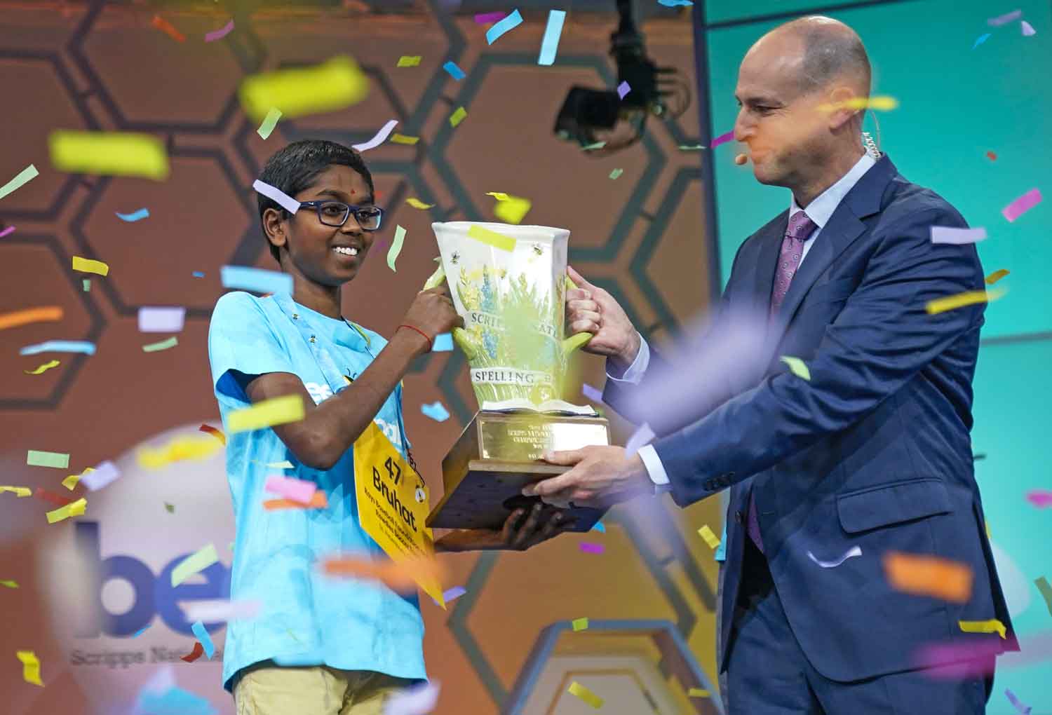 A man in a suit and headset gives Bruhat Soma a trophy as confetti flies through the air.