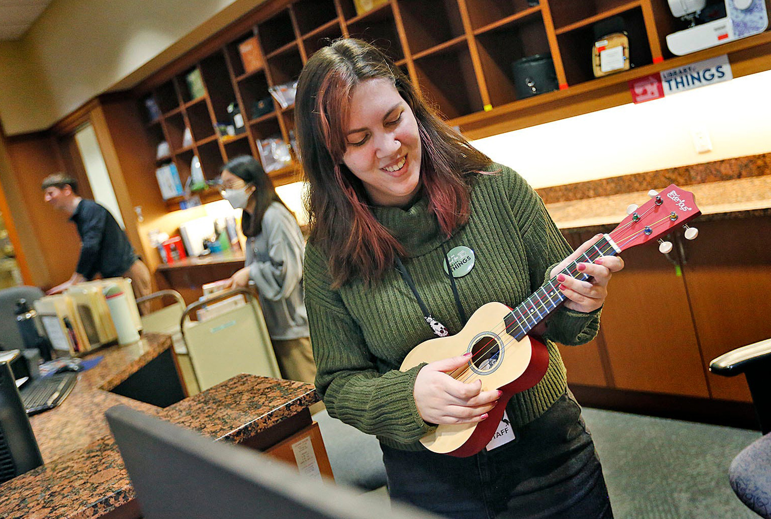 A librarian behind a circulation desk smiles as she plays a ukelele.