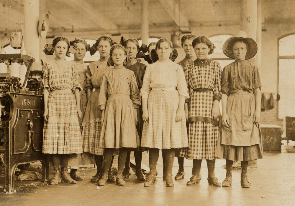 A group of girls pose in front of spinning machines in a factory.