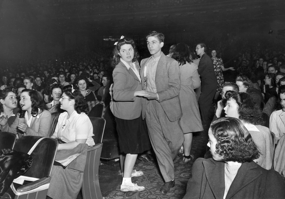 A teen couple dances in a theater as other young people are sitting or standing around them.