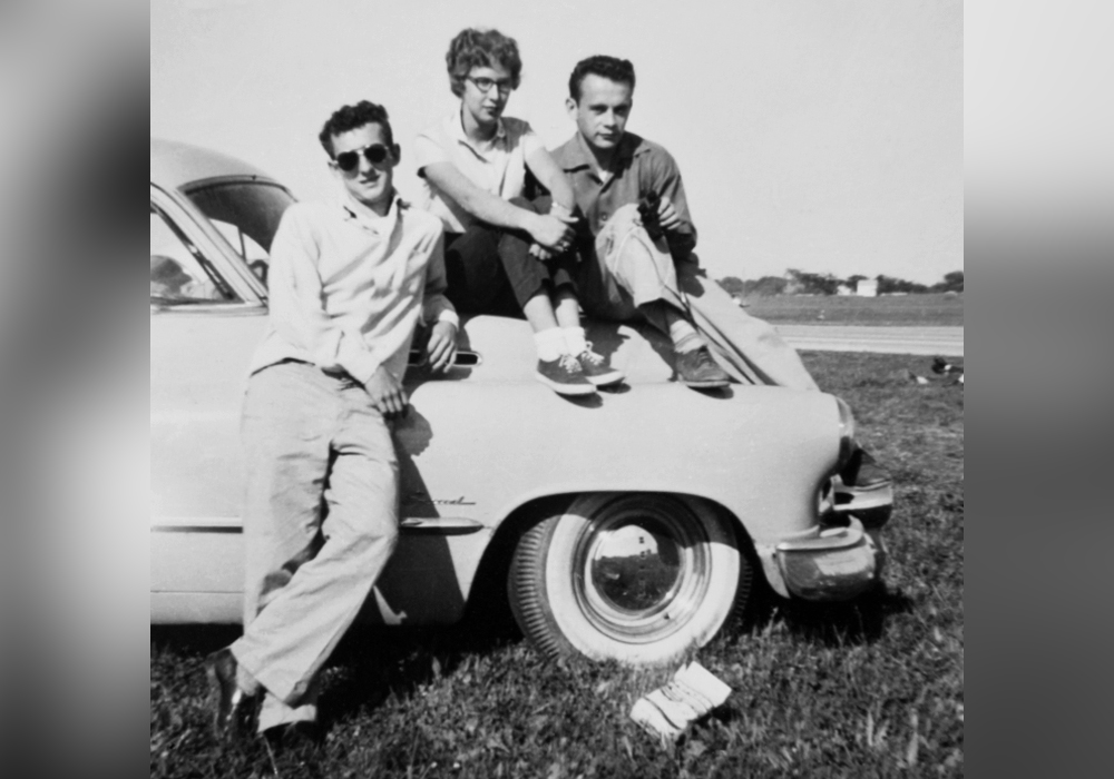 Three teenagers pose while sitting on or leaning against a 1950s car.